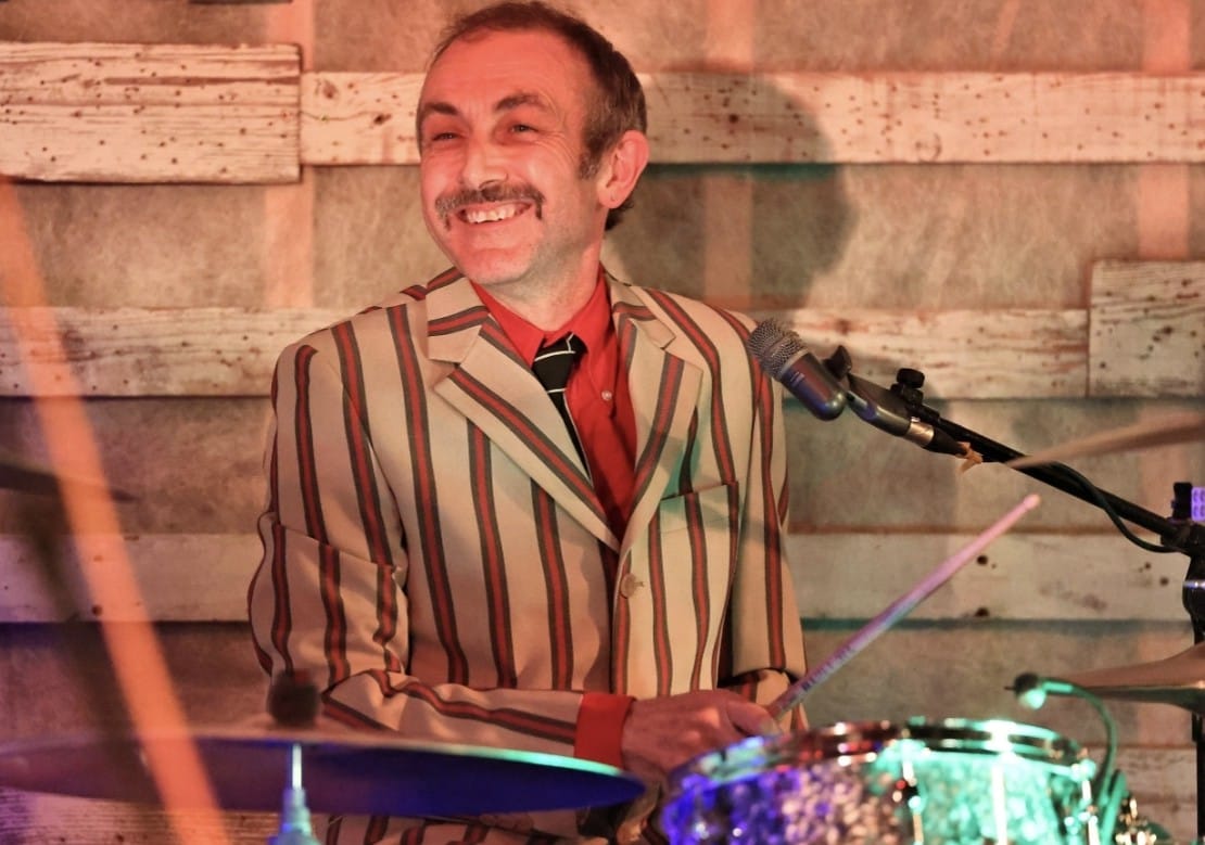 Spencer behind the drum kit in a striped jacket, smiling mid-song