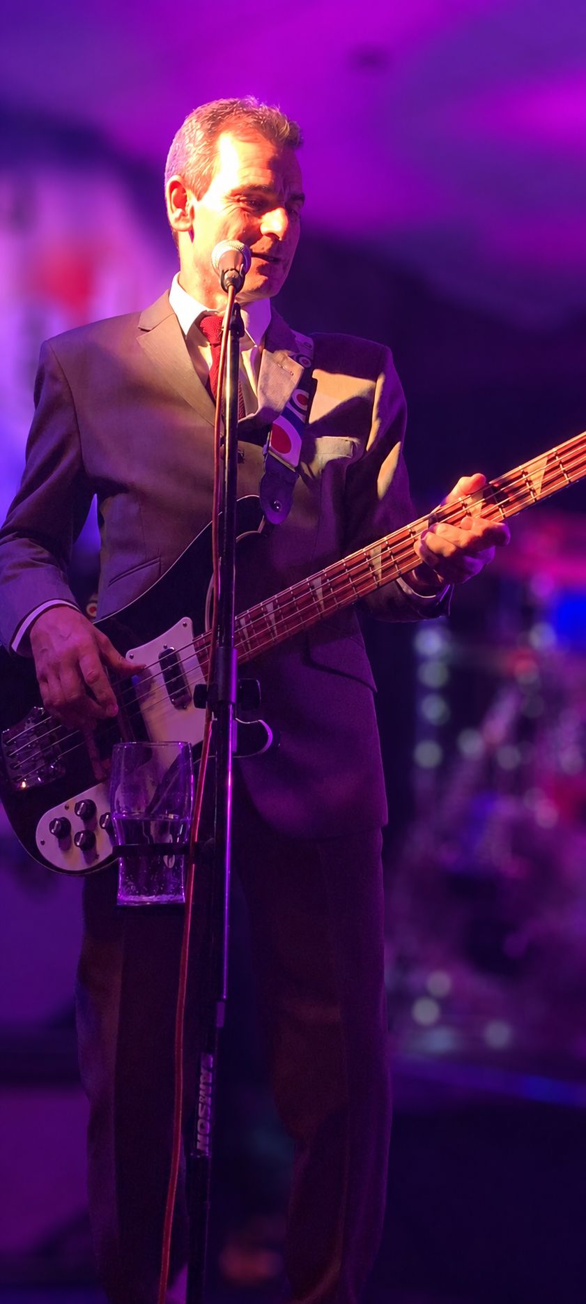 Bass player in a grey suit and red tie playing a Rickenbacker bass at the mic under purple stage lighting