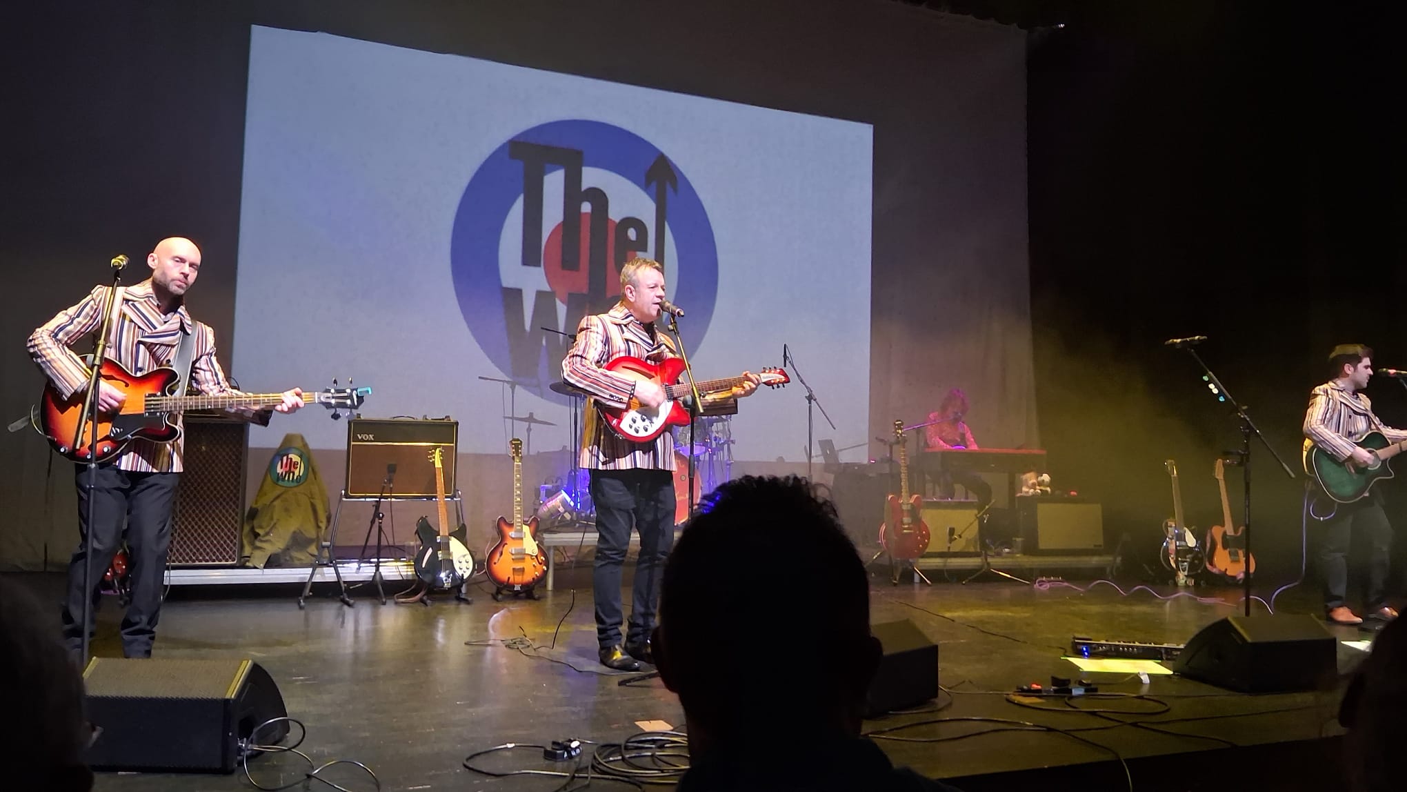 The band performing in front of a giant The Who target-logo backdrop, keys player at the back