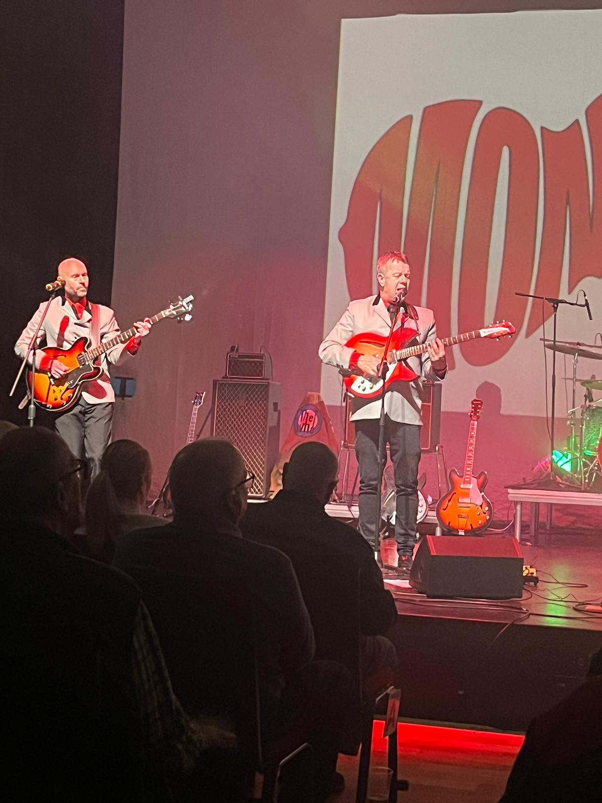 Two members of Back to the Sixties on guitar and vocals at their mics under red stage lighting