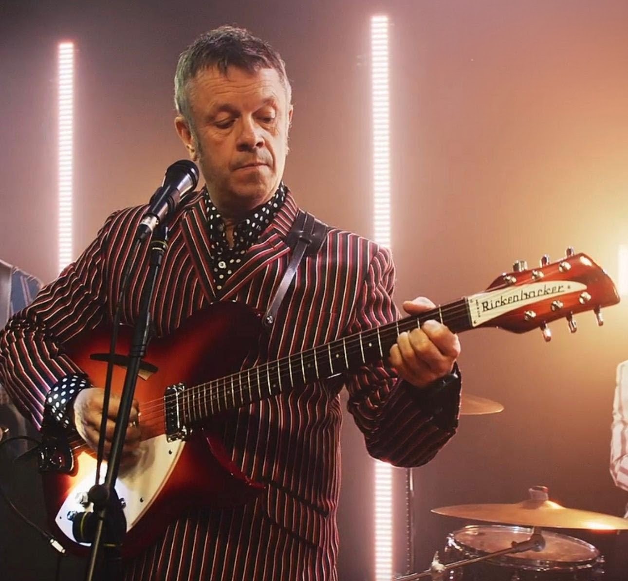 Gaz in a striped suit playing his Rickenbacker guitar at the mic