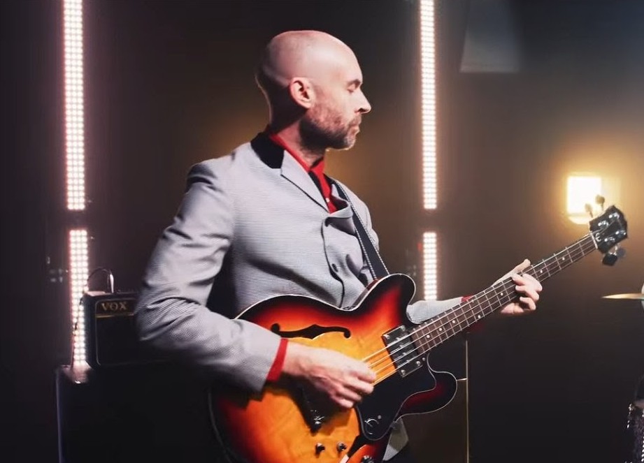 Phil on bass guitar in a grey suit beside a Vox amp under warm stage lighting